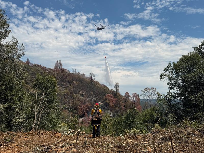 Helicopter with suspended bucket over a wooded hillside, firefighter in yellow foreground