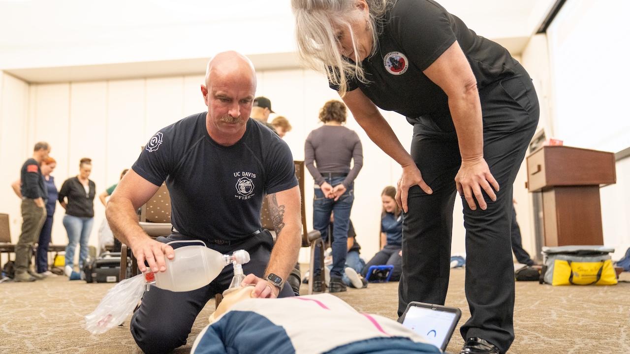 Two emergency responders kneel over a supine trainee during CPR training, AED monitor at side