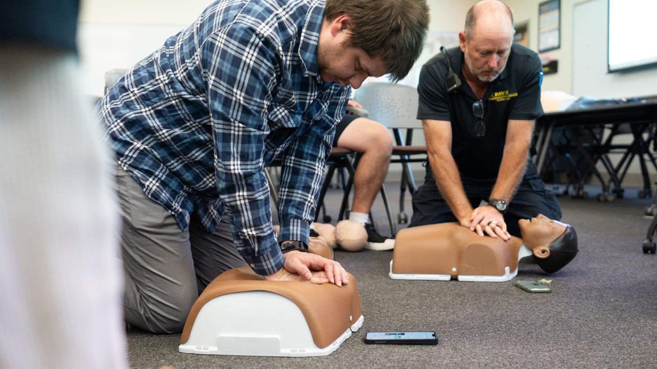 Two men kneeling practicing CPR on training mannequins in a classroom