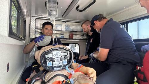 Two paramedic students sit in the back of an ambulance and supply oxygen to a manikin.